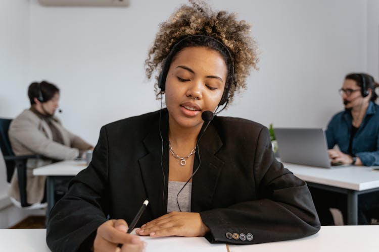 A Woman Wearing Black Blazer With Black Headset Holding A Pen