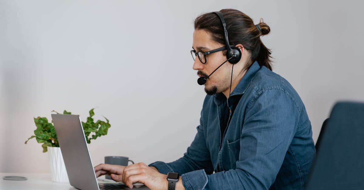 Adult man wearing a headset working on a laptop in an office space, providing remote customer support.