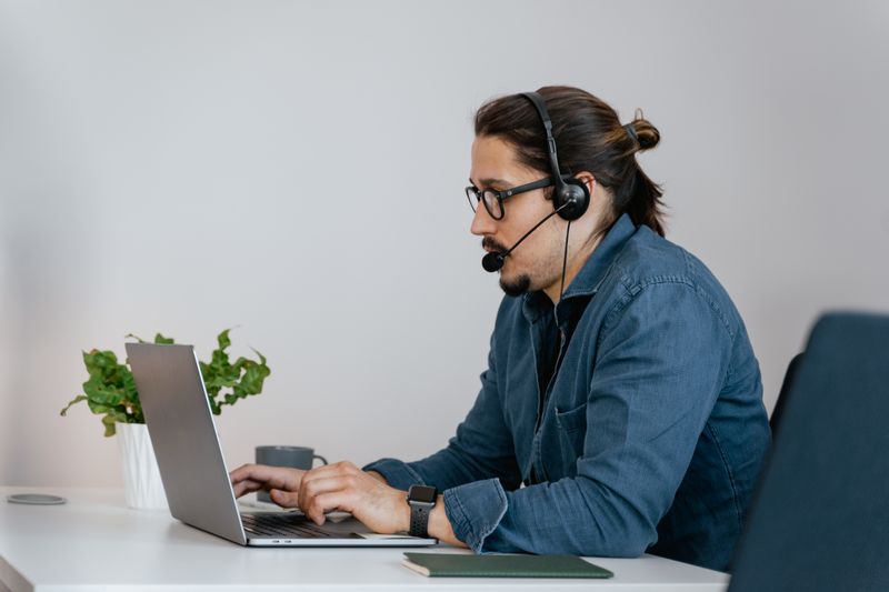 Adult man wearing a headset working on a laptop in an office space, providing remote customer support