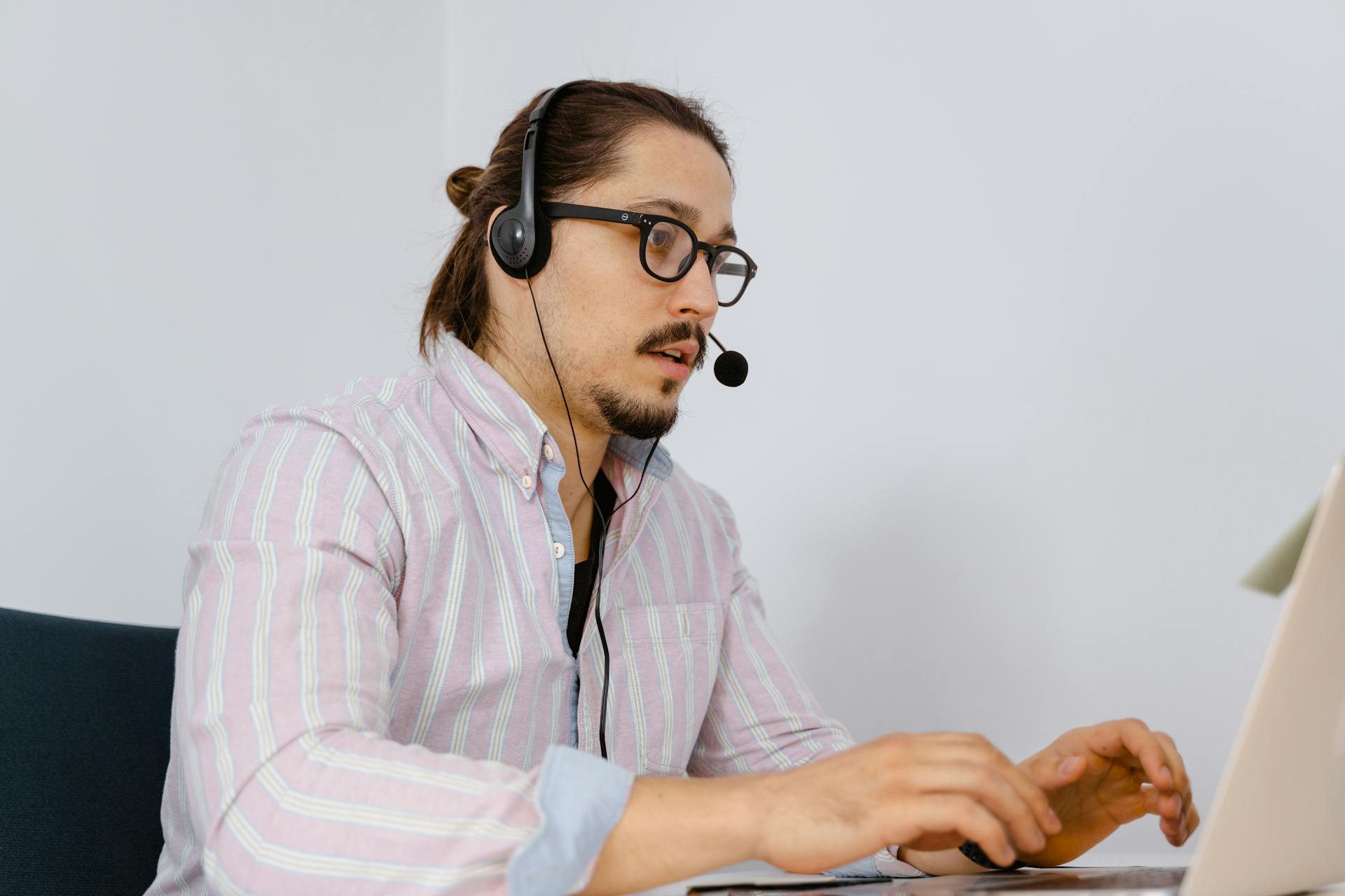 A young man in a striped shirt works at a laptop, wearing a headset for customer support.