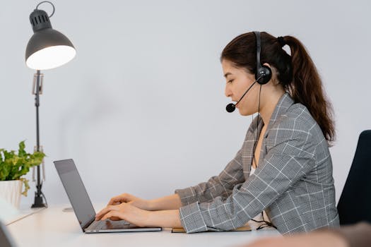 Proven AI Marketing Strategies for 2025 2 Professional woman in office with headset typing on laptop, asking ai virtual assistant support and ai marketing .