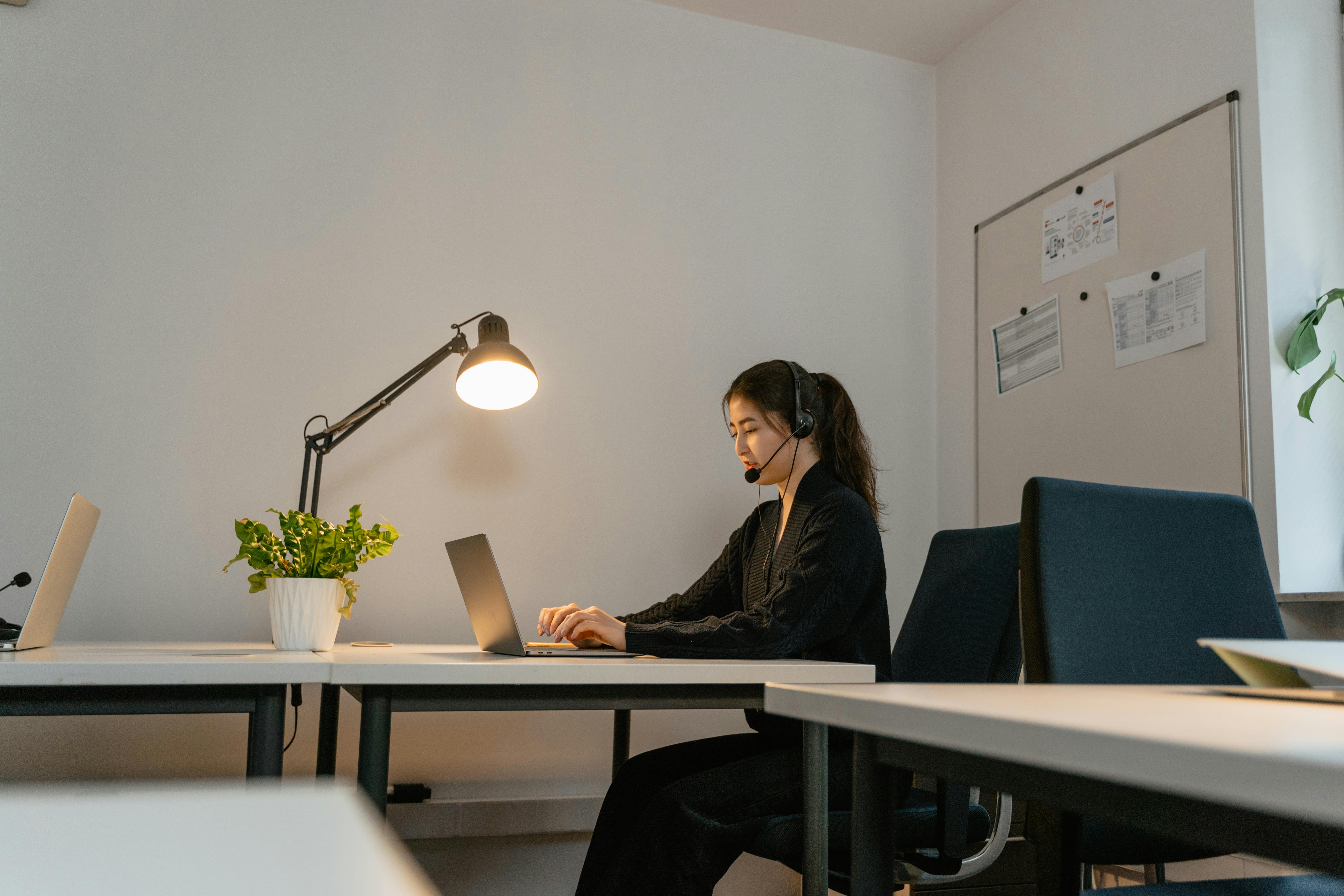 Free A woman in a modern office setting, wearing a headset and working on a laptop under a desk lamp. Stock Photo