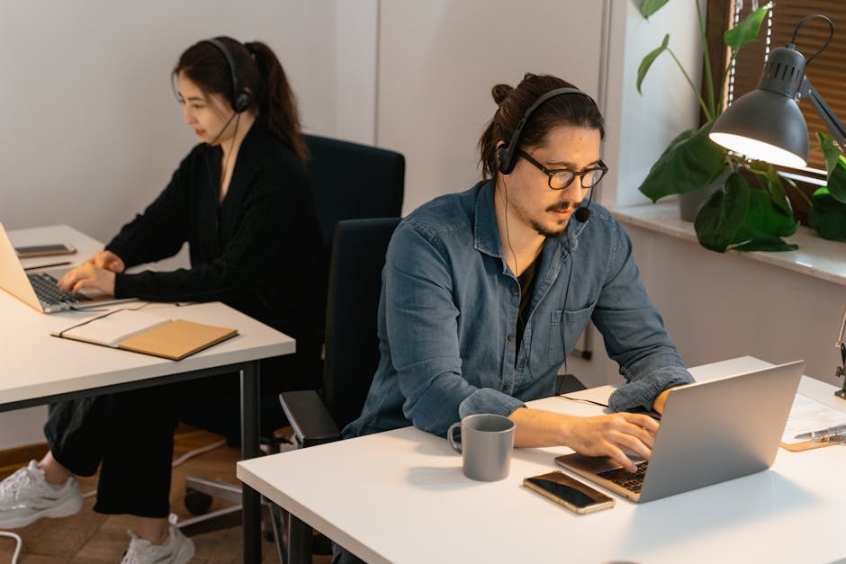 Two colleagues working in an office space, focused on laptops, wearing headsets for online communication.