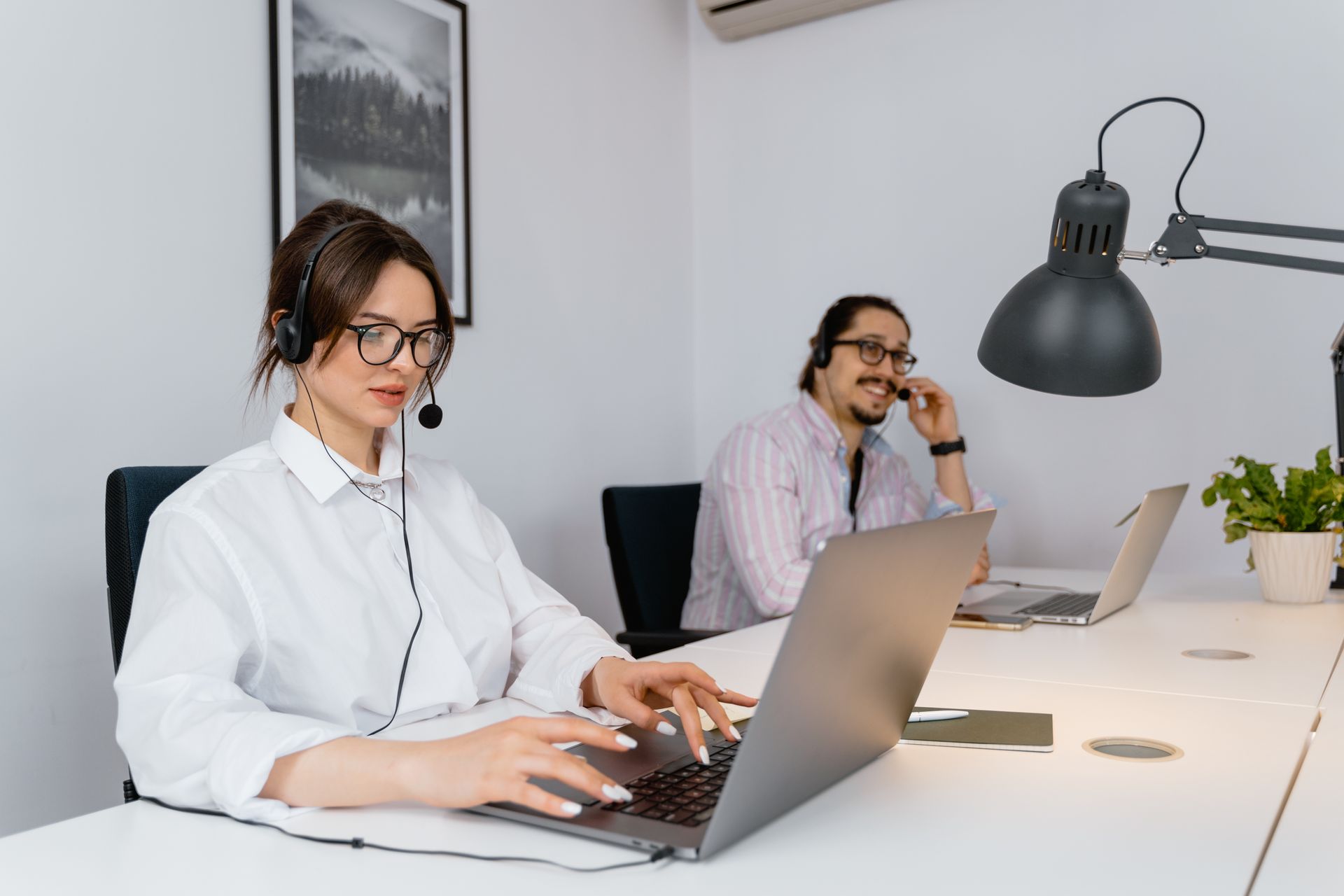 Two call center employees working with headsets and laptops in a modern office setting.