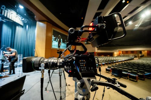 Modern professional video camera with lens placed on tripod near stage with unrecognizable male in concert hall