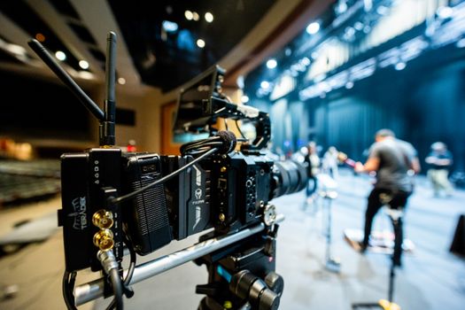 Modern video camera with lens on tripod recording unrecognizable people standing on stage in concert hall