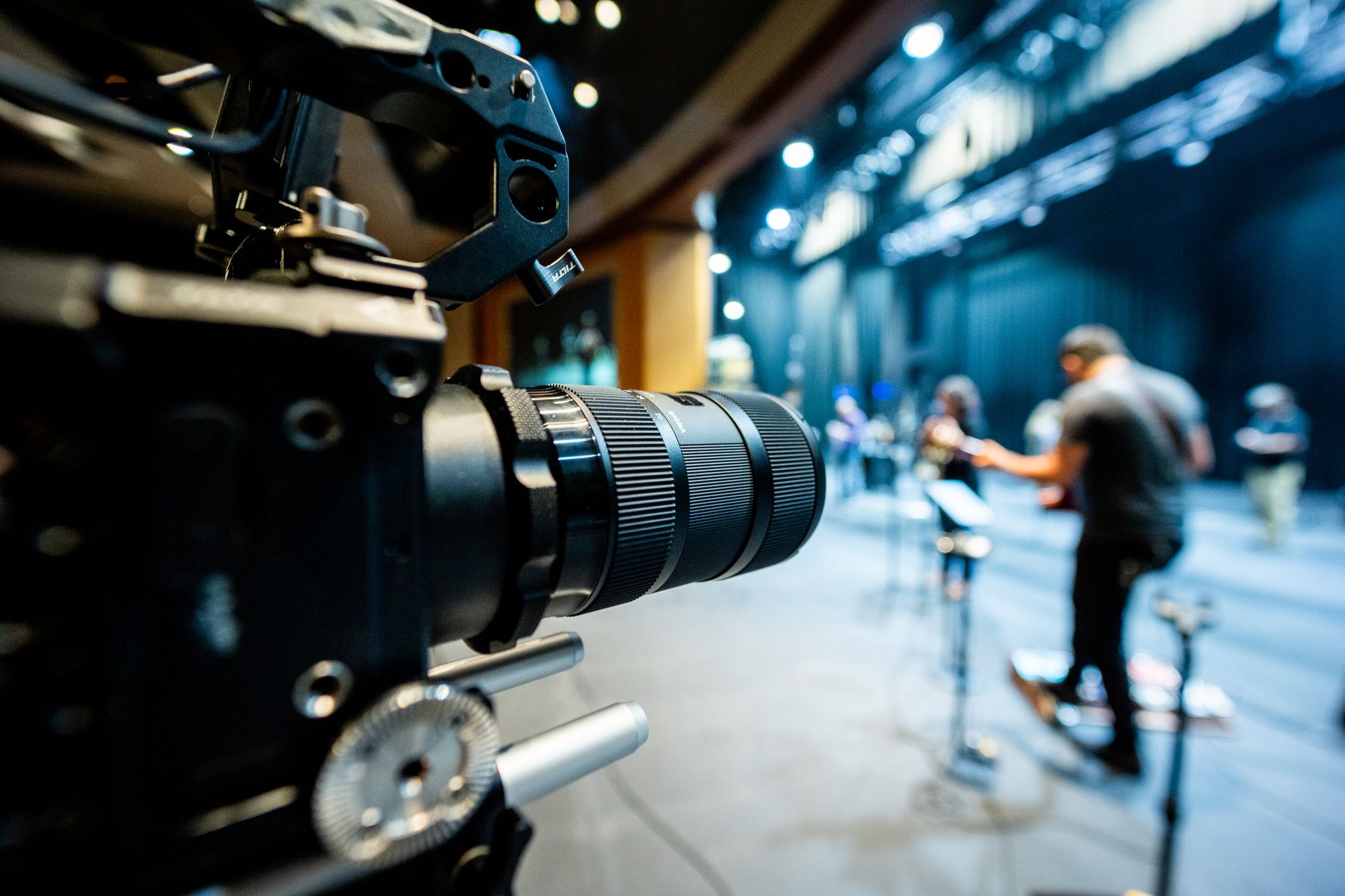 Closeup of professional video camera with lens recording unrecognizable people standing on stage illuminated by lights in studio