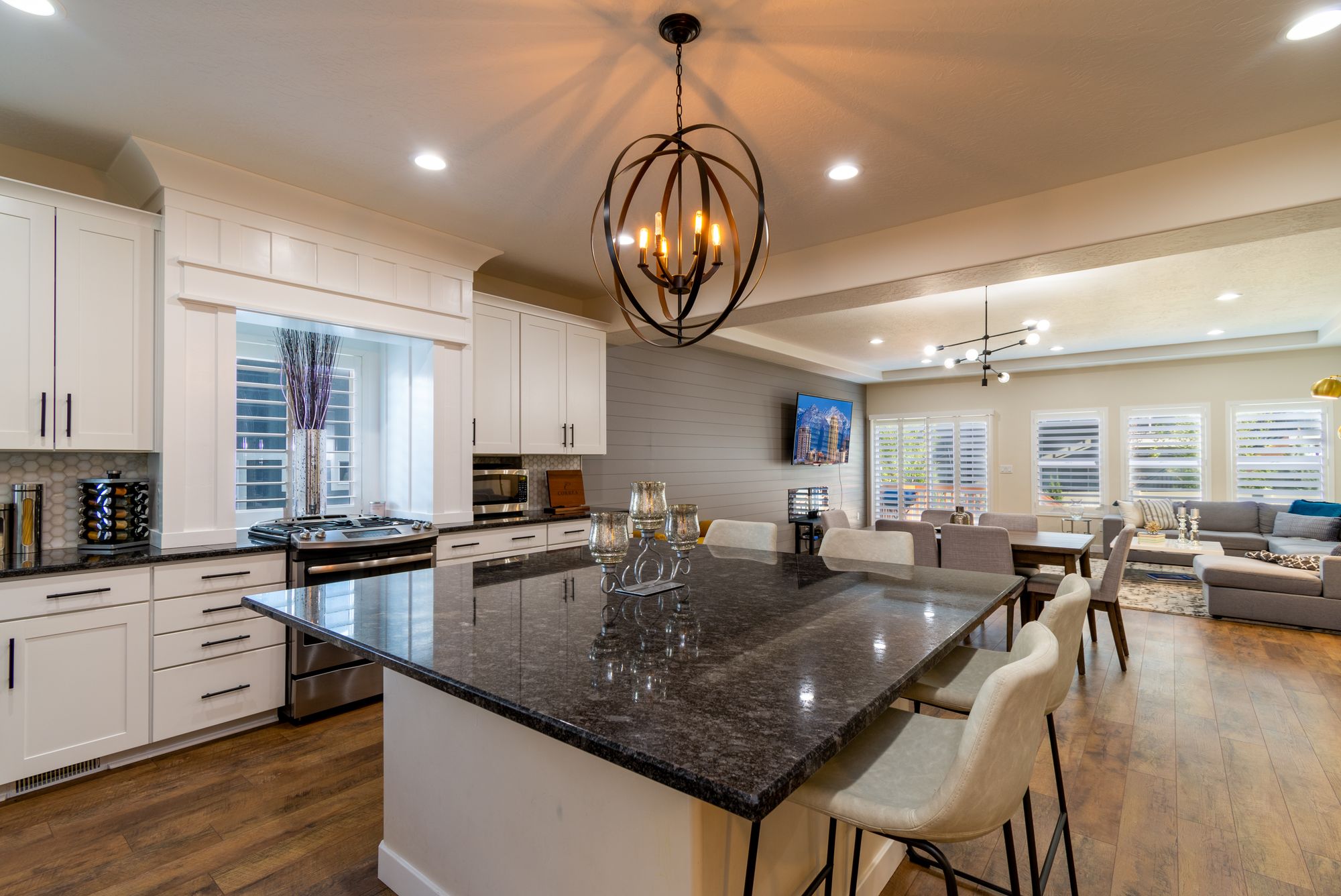 Open-plan kitchen and living area with white shaker cabinets, a large dark stone island, pendant lighting, dining seating, timber flooring, and adjoining lounge space.
