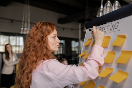 Red-haired woman writing on a whiteboard with sticky notes in a modern office setting.