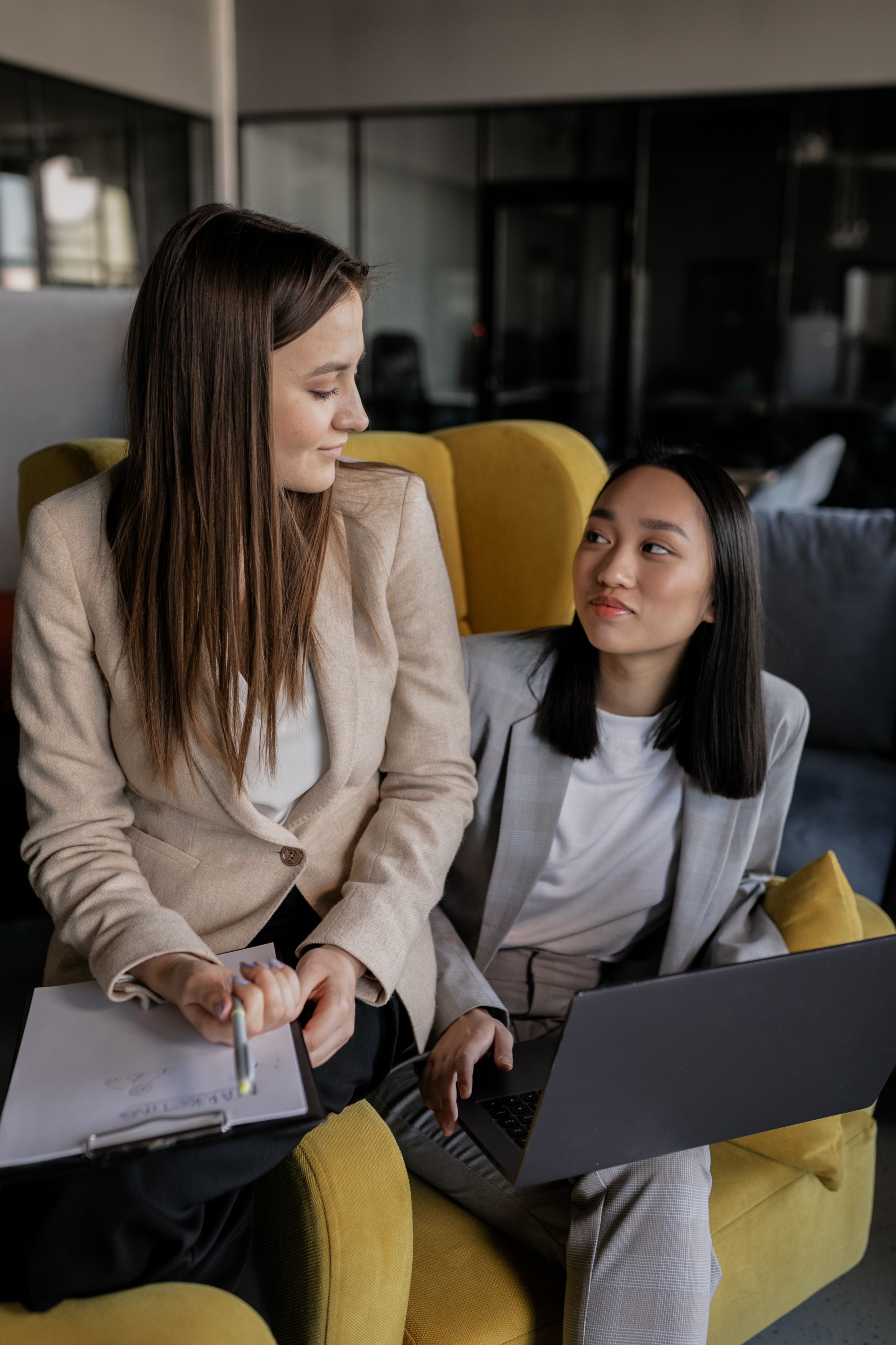 Two professionally dressed women engaged in discussion in an office lounge; one is holding a clipboard while the other works on a laptop.