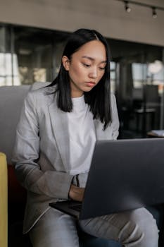 Focused Asian woman in business attire working on laptop in a modern office setting.