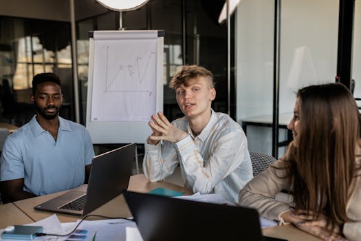 Three diverse colleagues engaged in a business strategy discussion in a modern office with a chart in the background.