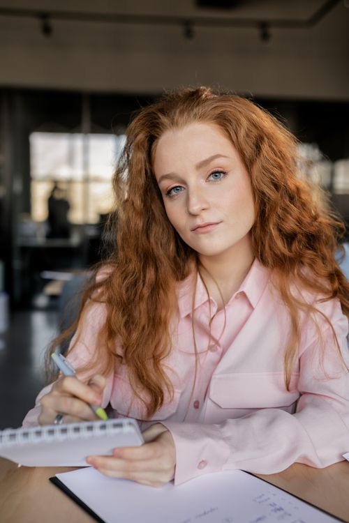 Free Red-haired woman writing at a desk in an indoor office environment. Stock Photo