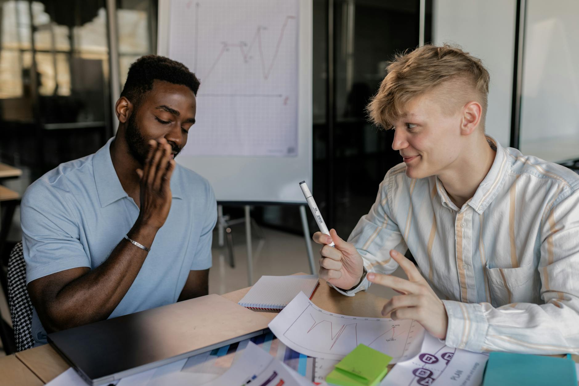 Two young men collaborating with charts, paper, and a laptop on a desk indoors.