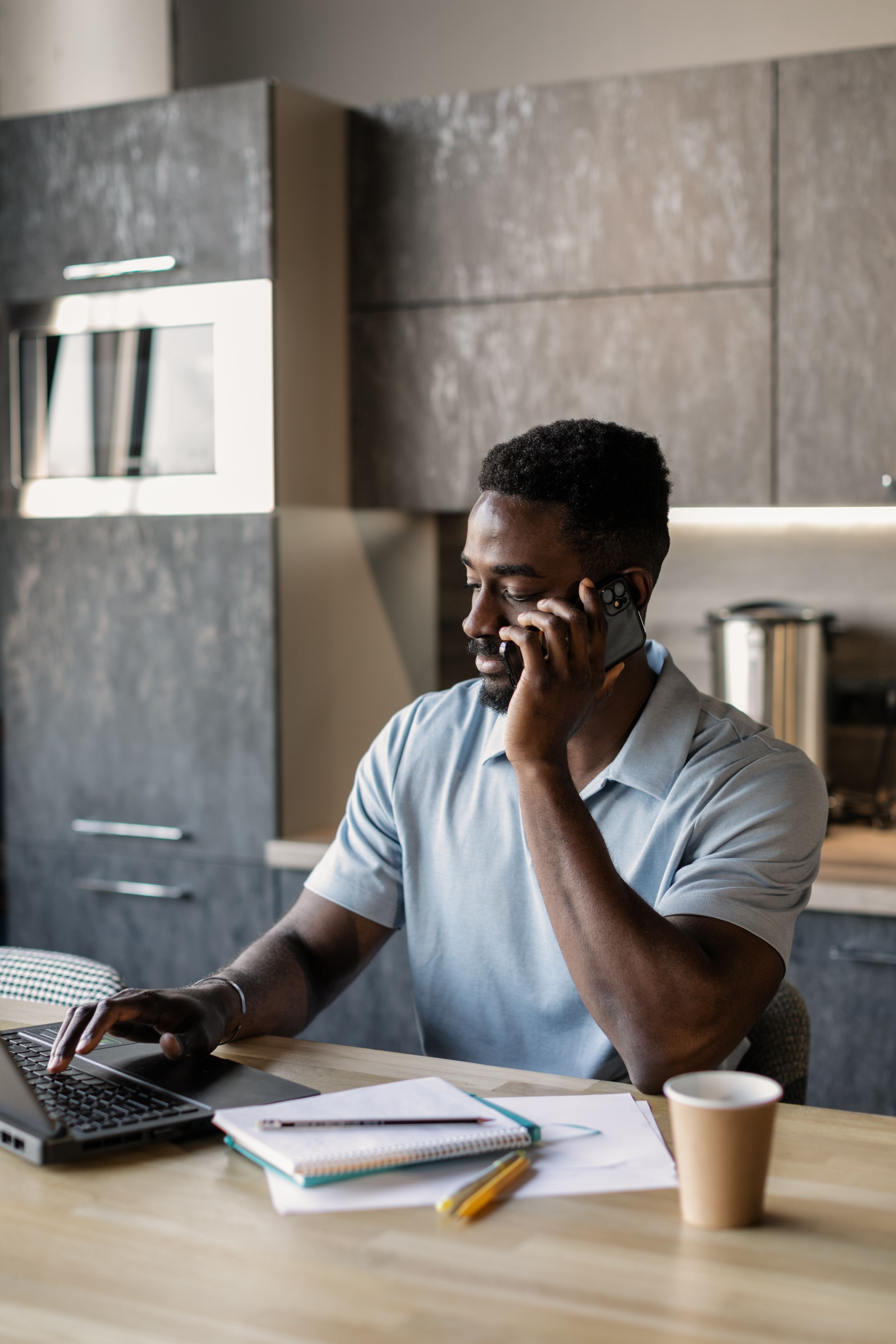 African American man talking on smartphone while typing on laptop, multitasking in a modern kitchen setting.