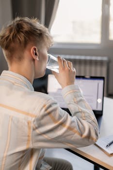 Young adult working from home takes a water break while using a laptop, creating a casual office setup.