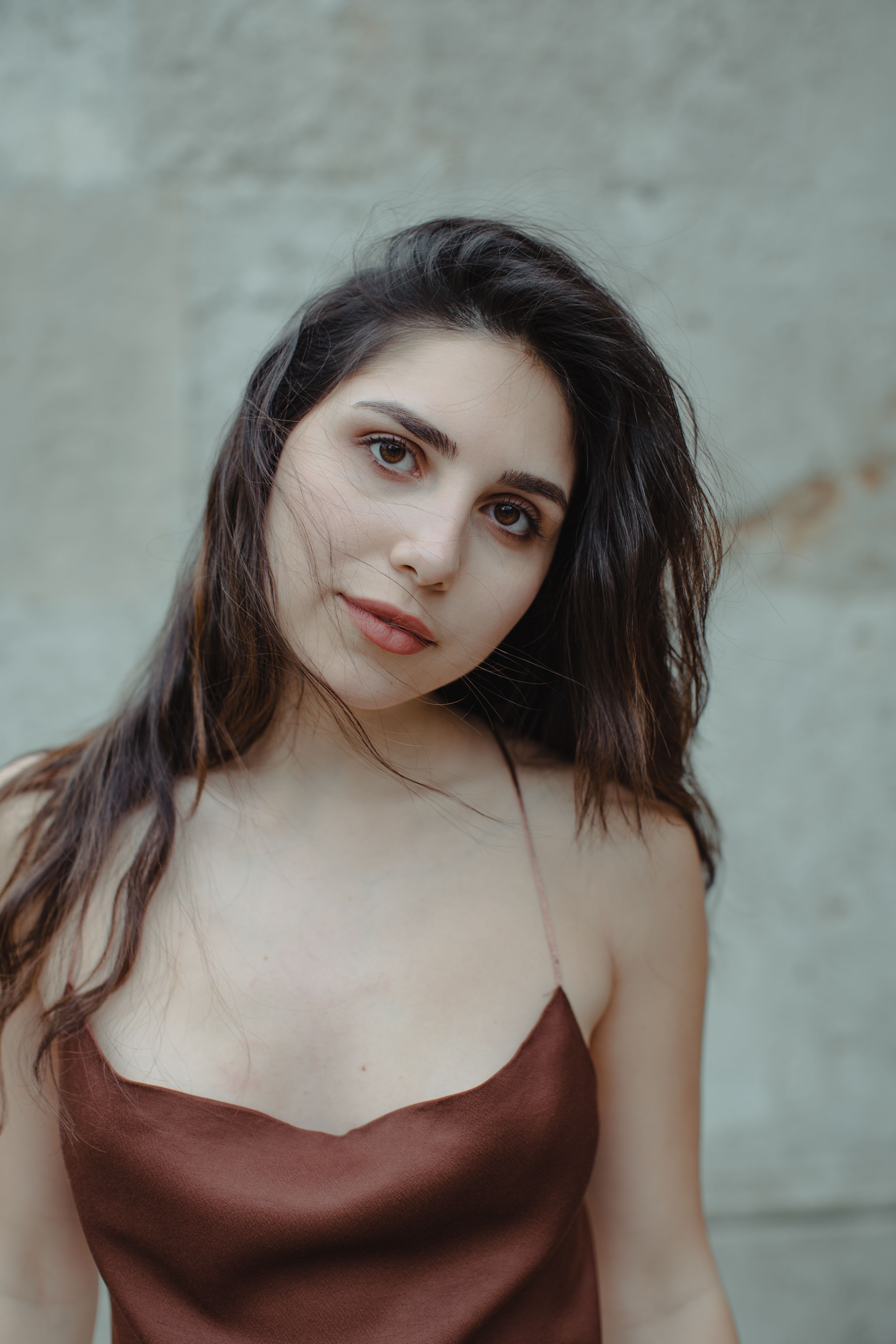 Charming calm female in brown strap top with long dark hair looking at camera against stone wall