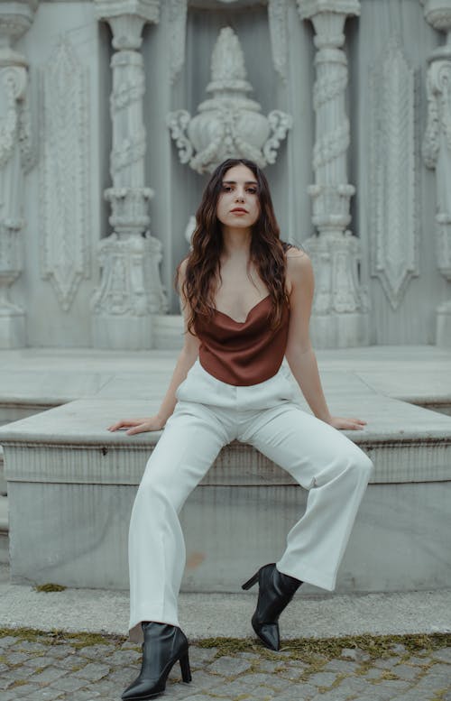 Confident stylish young female wearing trendy outfit and boots sitting on stone border and posing