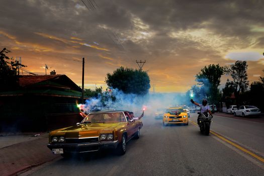 Vibrant wedding procession featuring vintage cars, motorcycles, and colorful smoke flares at sunset.