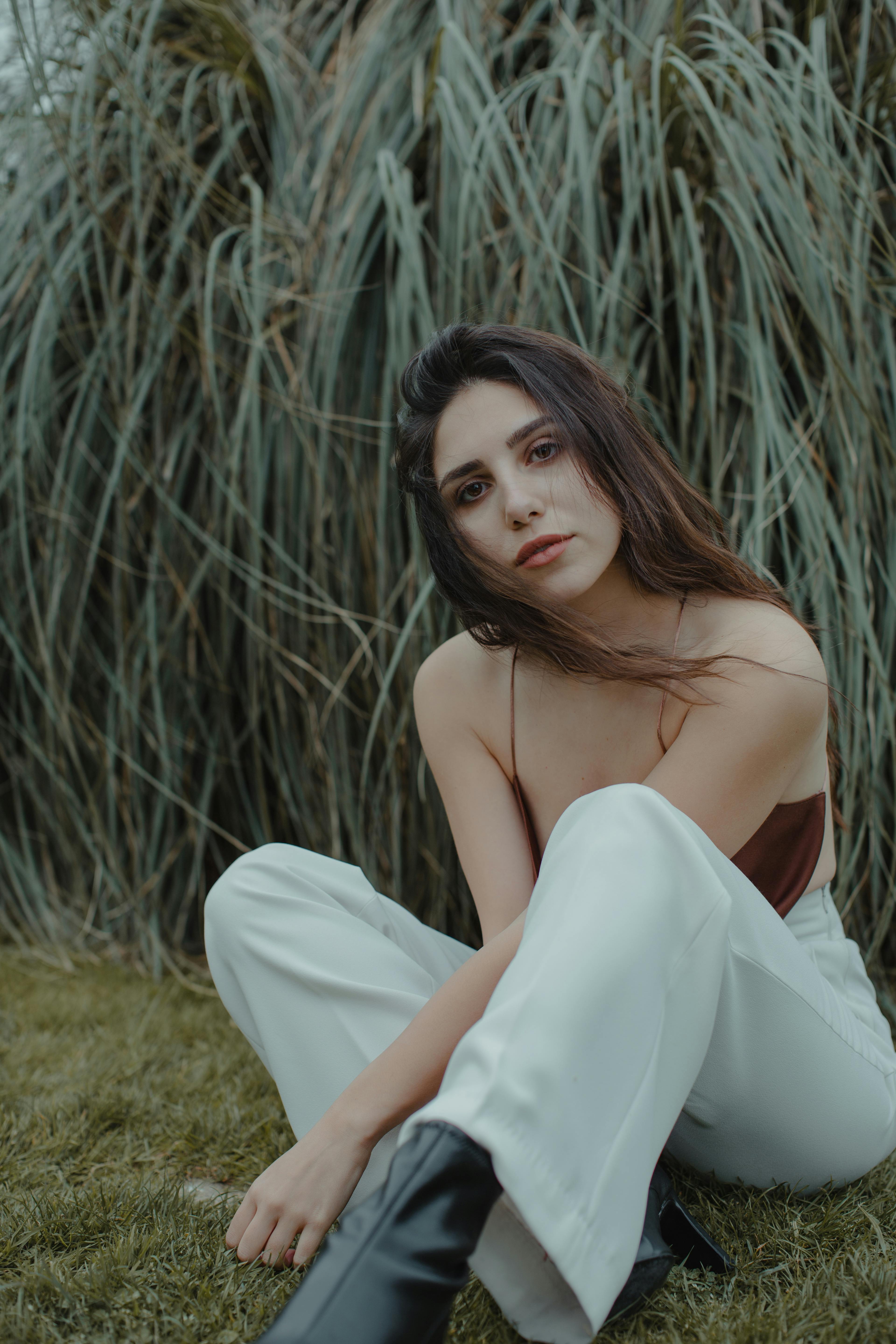 Confident brunette in casual clothes sitting on grass near plants and looking at camera