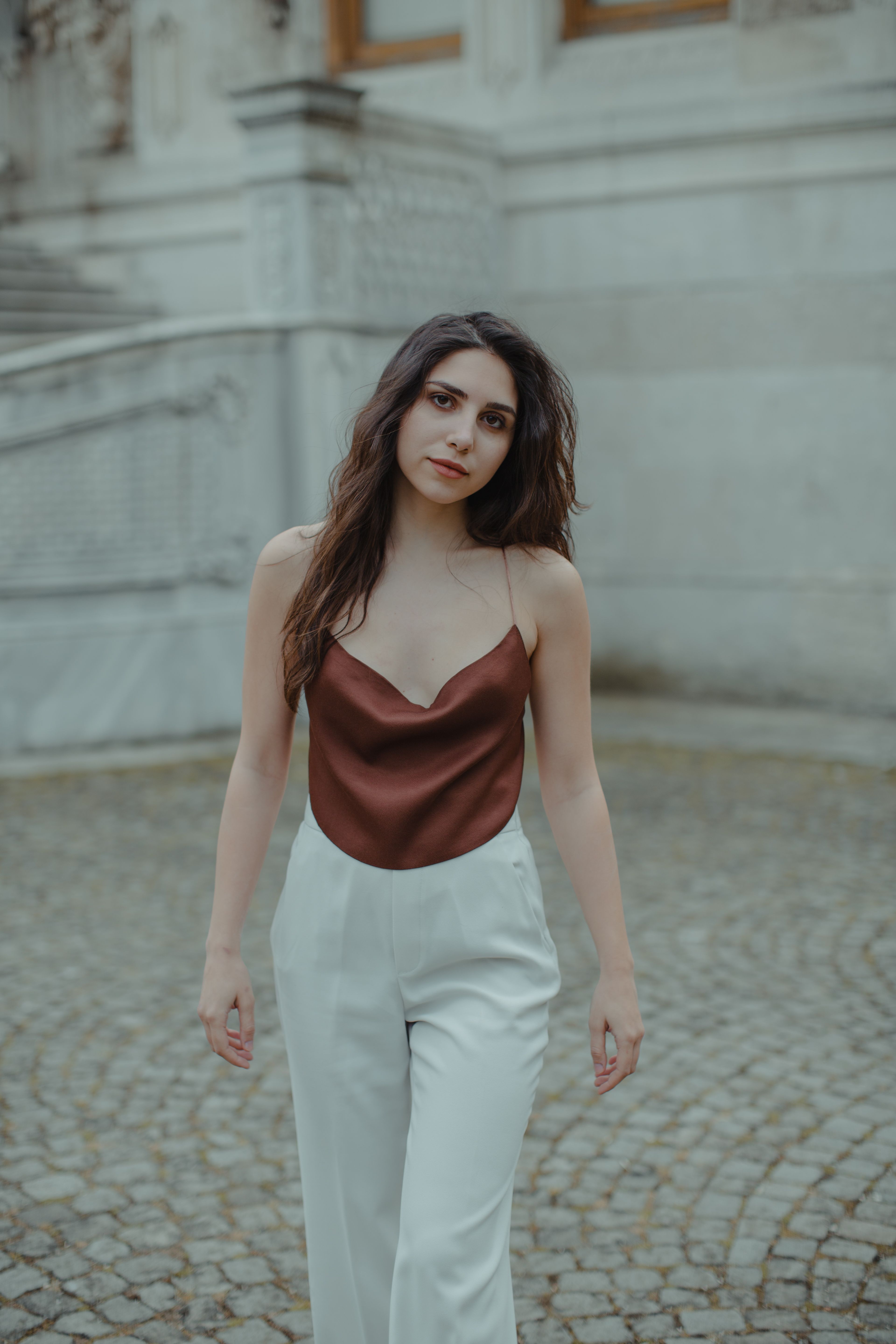 An elegant woman in a silky top and white pants walks confidently outdoors.