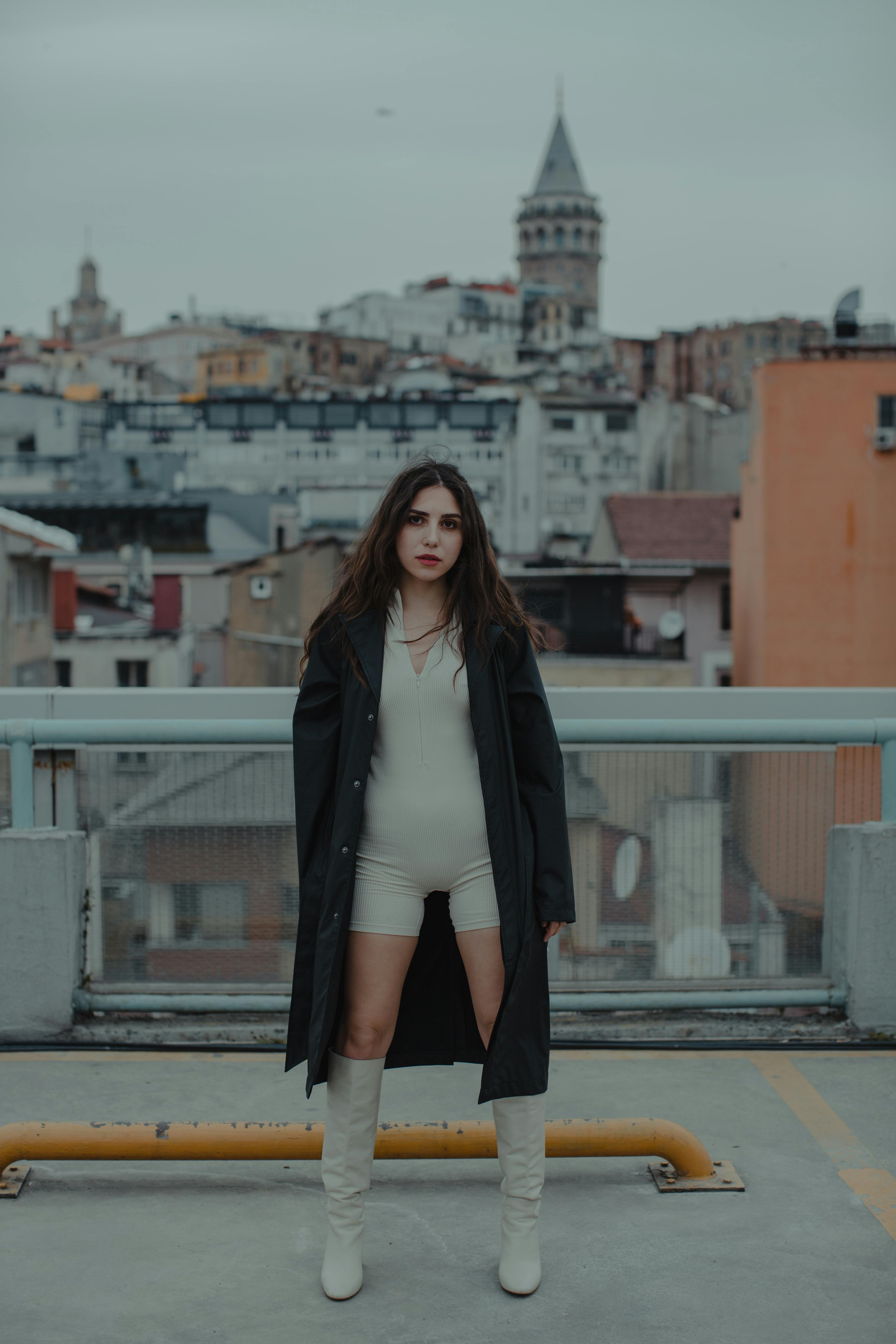 Fashionable woman in coat and boots stands confidently in an urban area with a historic tower in the background.
