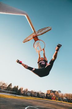 Full body of faceless energetic sportsman in activewear hanging on basketball hoop with legs apart during training on sports ground in city