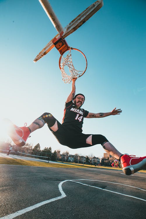 Full body of active male player in sportswear hanging on basketball hoop while training on sunny sports ground in city