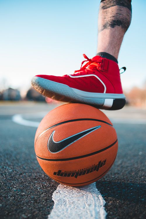 Free Crop faceless sportive man in red sneakers with foot on orange basketball placed on asphalt sports ground during training on street Stock Photo