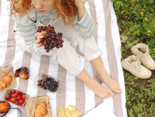 Free Young woman with curly hair holding grapes at a picnic surrounded by fruits and snacks. Stock Photo