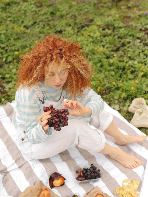 Free Woman with Curly Hair Sitting on a Picnic Blanket while Looking at the Grapes she is Holding Stock Photo
