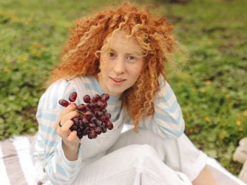 A Woman in Curly Hair Holding Grapes while Smiling at the Camera