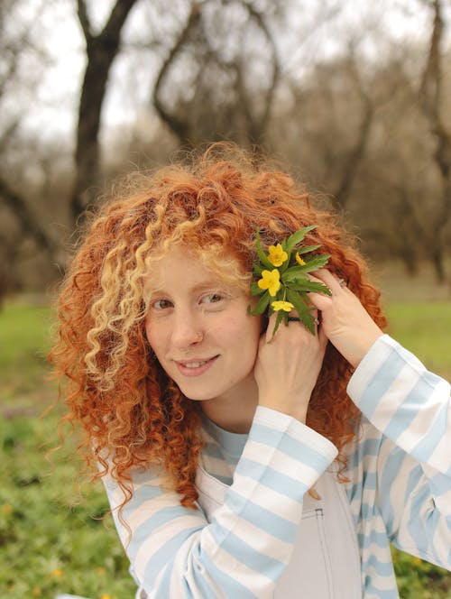 Woman in Curly Hair Clipping the Flowers with Green Leaves on Her Ear while Smiling at the Camera