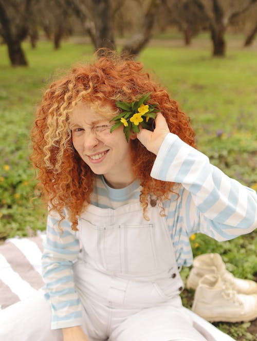 Woman in Curly Hair Clipping the Flowers with Green Leaves on Her Ear while Smiling at the Camera