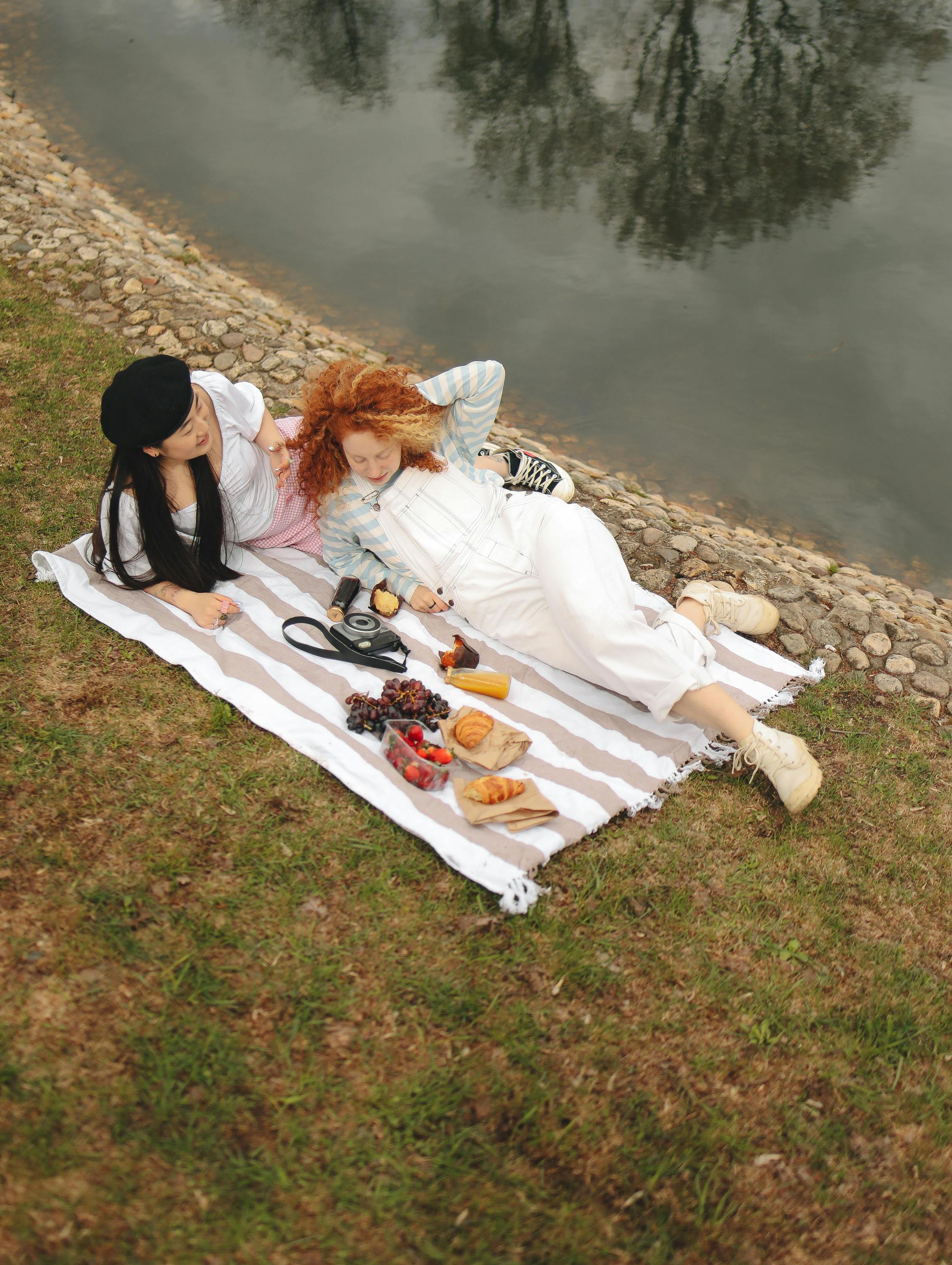 Two women enjoying a relaxing picnic by the lakeside with food and camera.