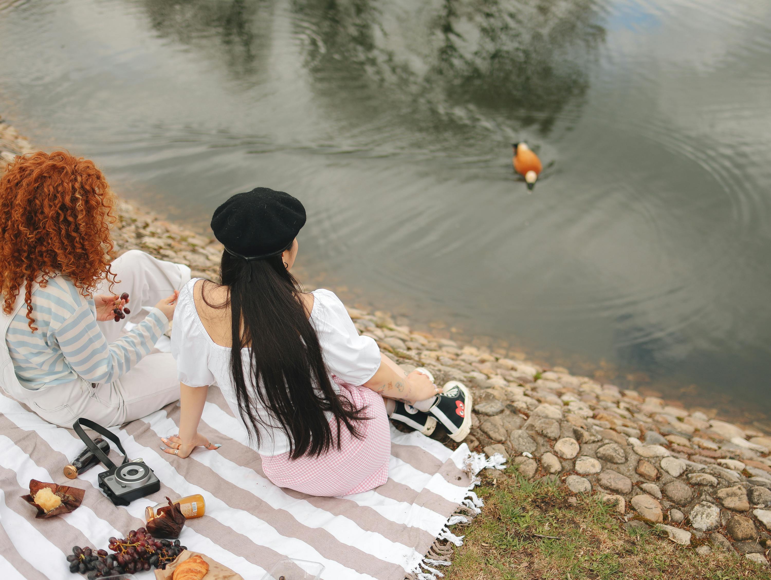 Two young women enjoying an outdoor picnic by a lake with a calm duck swimming nearby.
