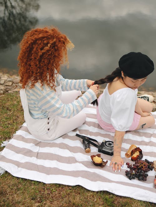 Woman in Curly Hair Braiding the Hair of the Woman in White Blouse while Sitting on a Picnic Blanket Near the Lake