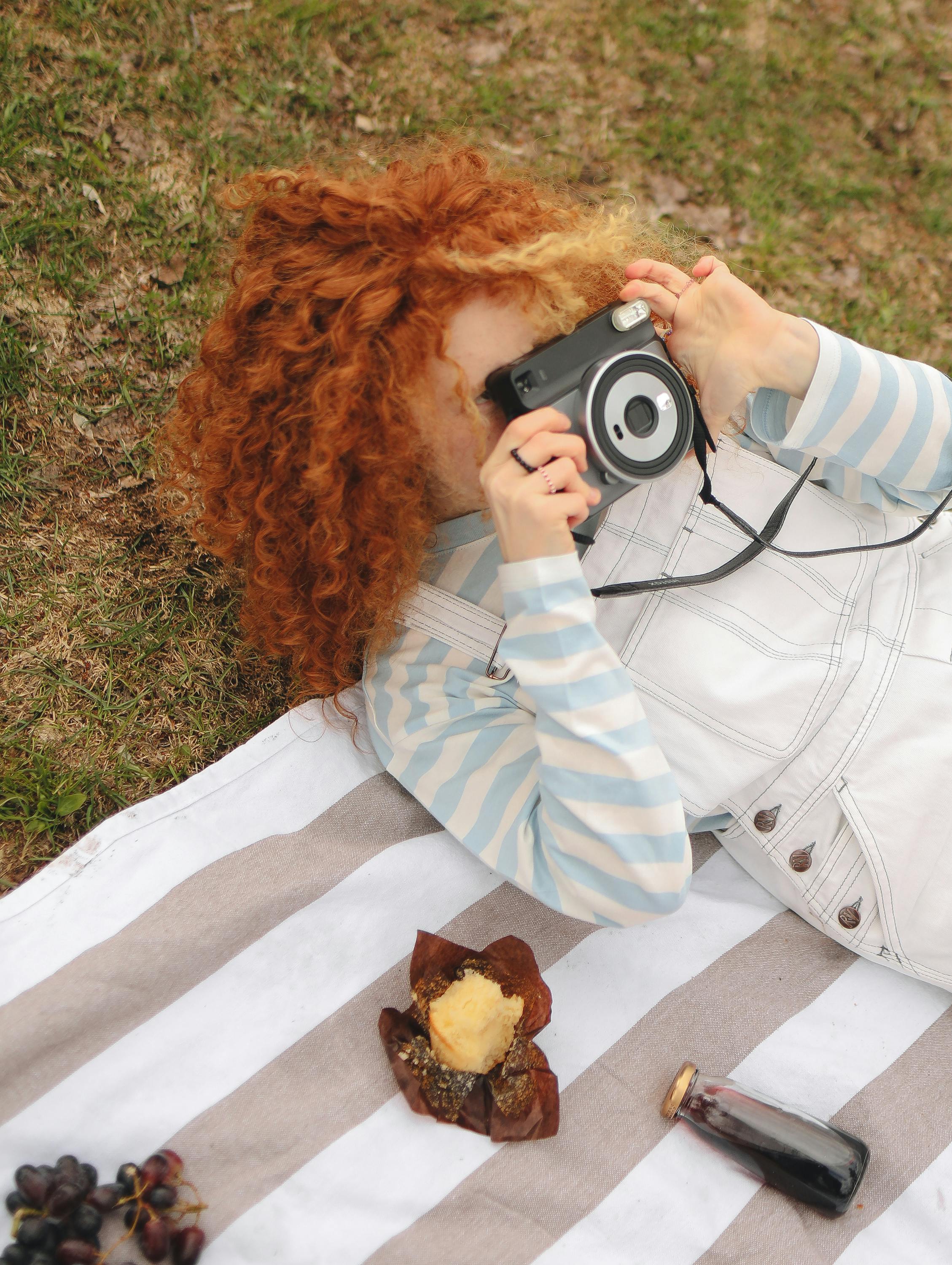 A red-haired woman enjoying a sunny day, capturing moments with her instant camera during a picnic.