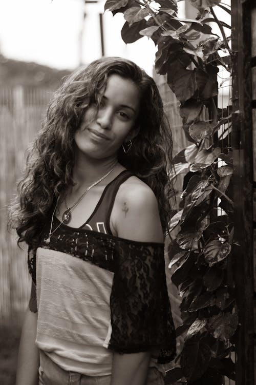 Free Grayscale Photo of a Woman with Curly Hair Standing Near Plants while Smiling at the Camera Stock Photo