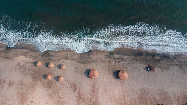 Drone shot capturing sandy beach with waves and straw beach huts from above.