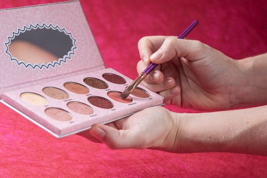 Close-up of hands holding and using an eye shadow palette with makeup brush on pink background.