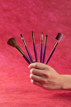 A hand showcasing a variety of makeup brushes against a bold red background.