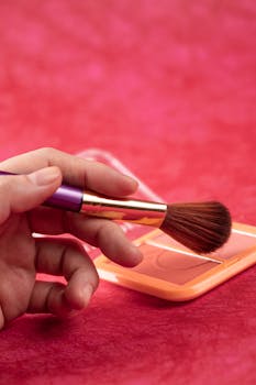 A hand holding a makeup brush over a blush palette on a vibrant background.