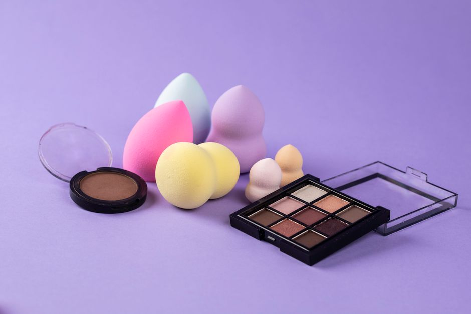 Aesthetic shot of makeup sponges and eyeshadow on purple backdrop.