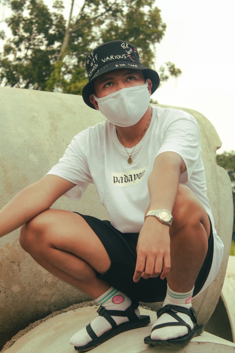 Man Wearing Bucket Hat And Face Mask Sitting On Concrete Pipes