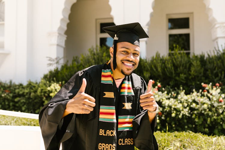 A Smiling Man In Black Academic Regalia Doing Thumbs Up