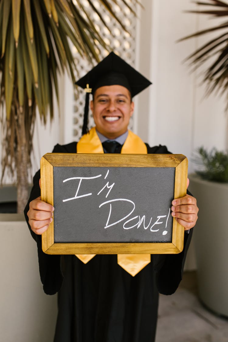 A Man Wearing Black Toga And Graduation Cap Holding A Small Board While Smiling At The Camera