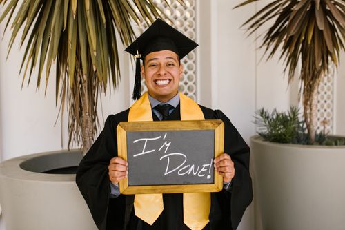 Free Happy graduate holding chalkboard outside, celebrating achievement in cap and gown. Stock Photo