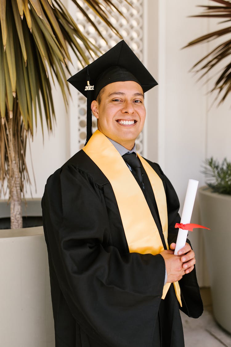 A Man Wearing Black Toga Holding Rolled Certificate While Smiling At The Camera