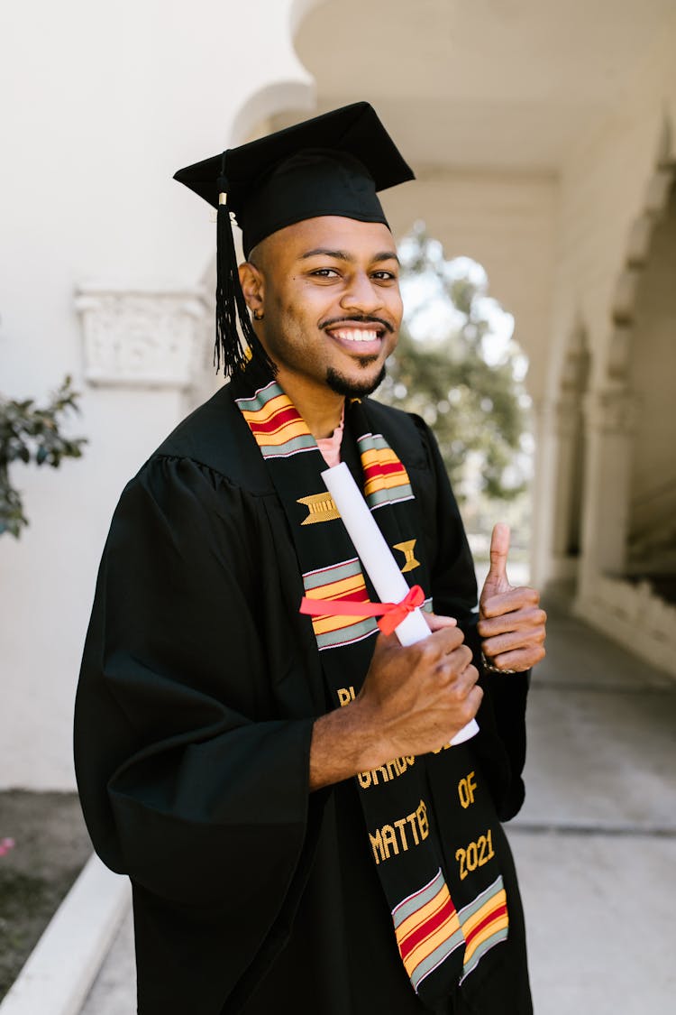 Man In Black Academic Dress Holding A Rolled Certificate While Smiling At The Camera